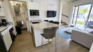 Eine kompakte Küche mit integriertem Mikrowellenherd, Kaffeemaschine und Spüle. Ein Essbereich mit Tisch und Stühlen lädt zum Verweilen ein, und im Wohnbereich befindet sich eine Couch. Ein Fernseher ist an der Wand befestigt, und ein Schild trägt die Aufschrift 'SURF SHACK'.