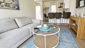 Ein modernes Wohnzimmer mit einem grauen Sofa, einem Glastisch mit blauem Tablett und einem im Hintergrund sichtbaren Küchenbereich.