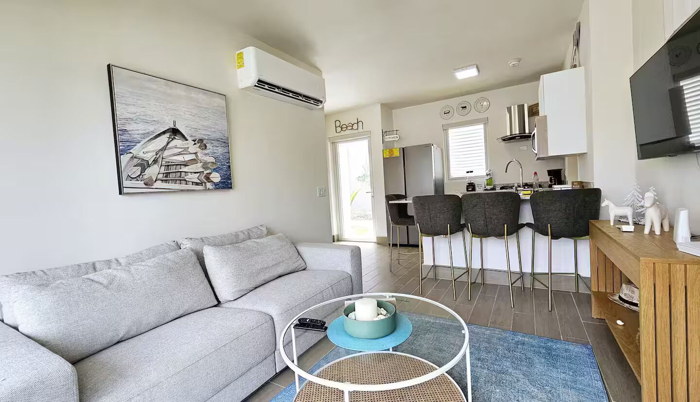 Een moderne woonkamer met een grijze bank, een salontafel en een eethoek met barstoelen.