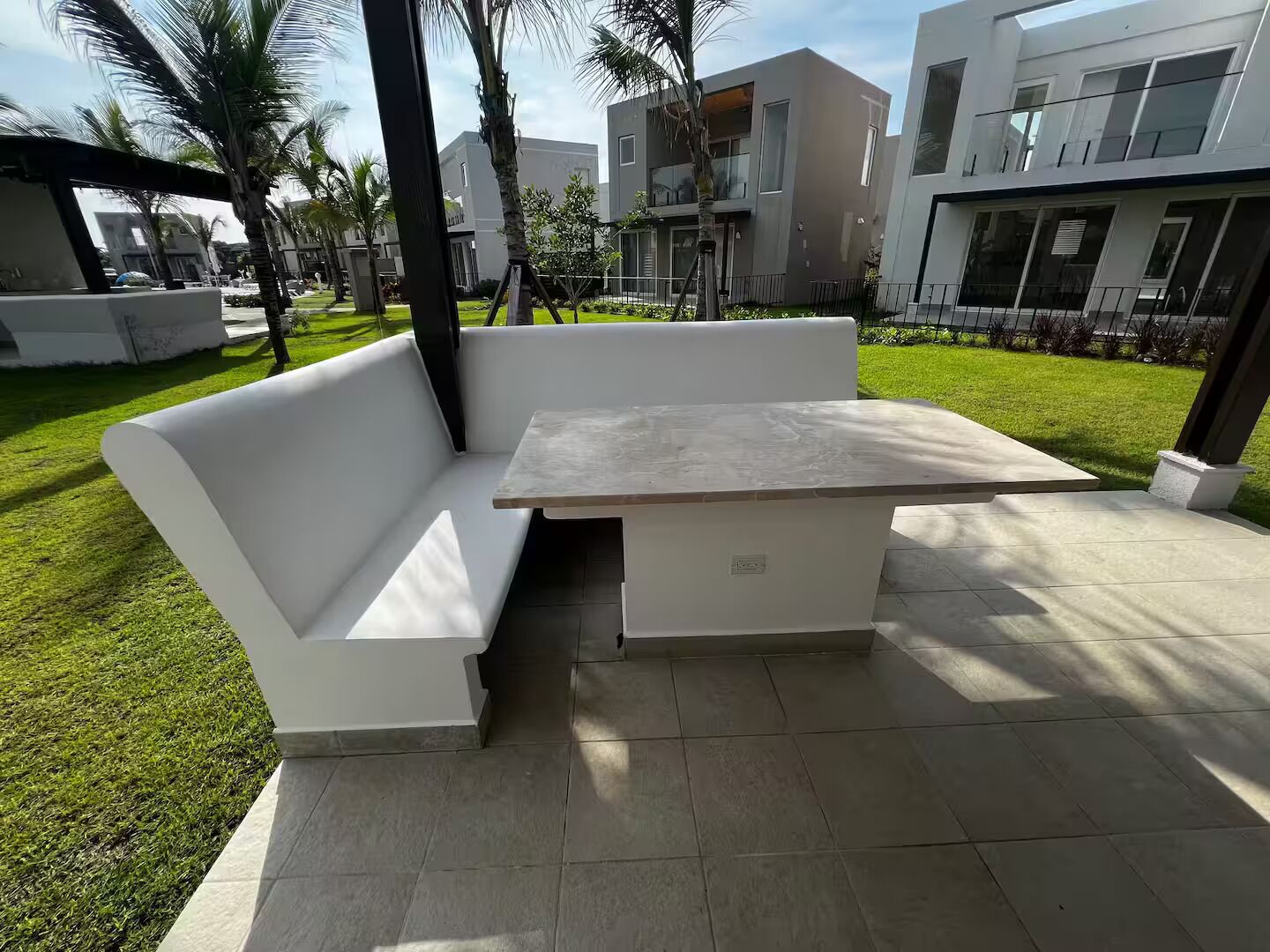 Uma área de estar moderna ao ar livre, com um sofá branco e uma mesa, cercada por um gramado bem cuidado e palmeiras, com edifícios contemporâneos ao fundo.