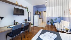 Ein modernes Hotelzimmer mit Essbereich, einem an der Wand befestigten Fernseher, einem Bett mit ordentlich gefalteten Handtüchern und einer kleinen Küchenzeile.