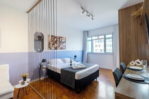Une chambre d’hôtel avec un lit, une table à manger avec des chaises, une fenêtre donnant sur des immeubles et un rideau.