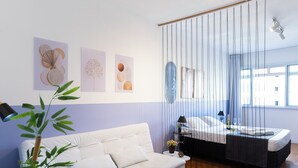 Ein modernes Zimmer mit einem weißen Sofa, einem Bett mit schwarzem Kopfteil und einem Fenster mit Vorhängen. Auf der linken Seite befinden sich Wandbilder und eine Pflanze.