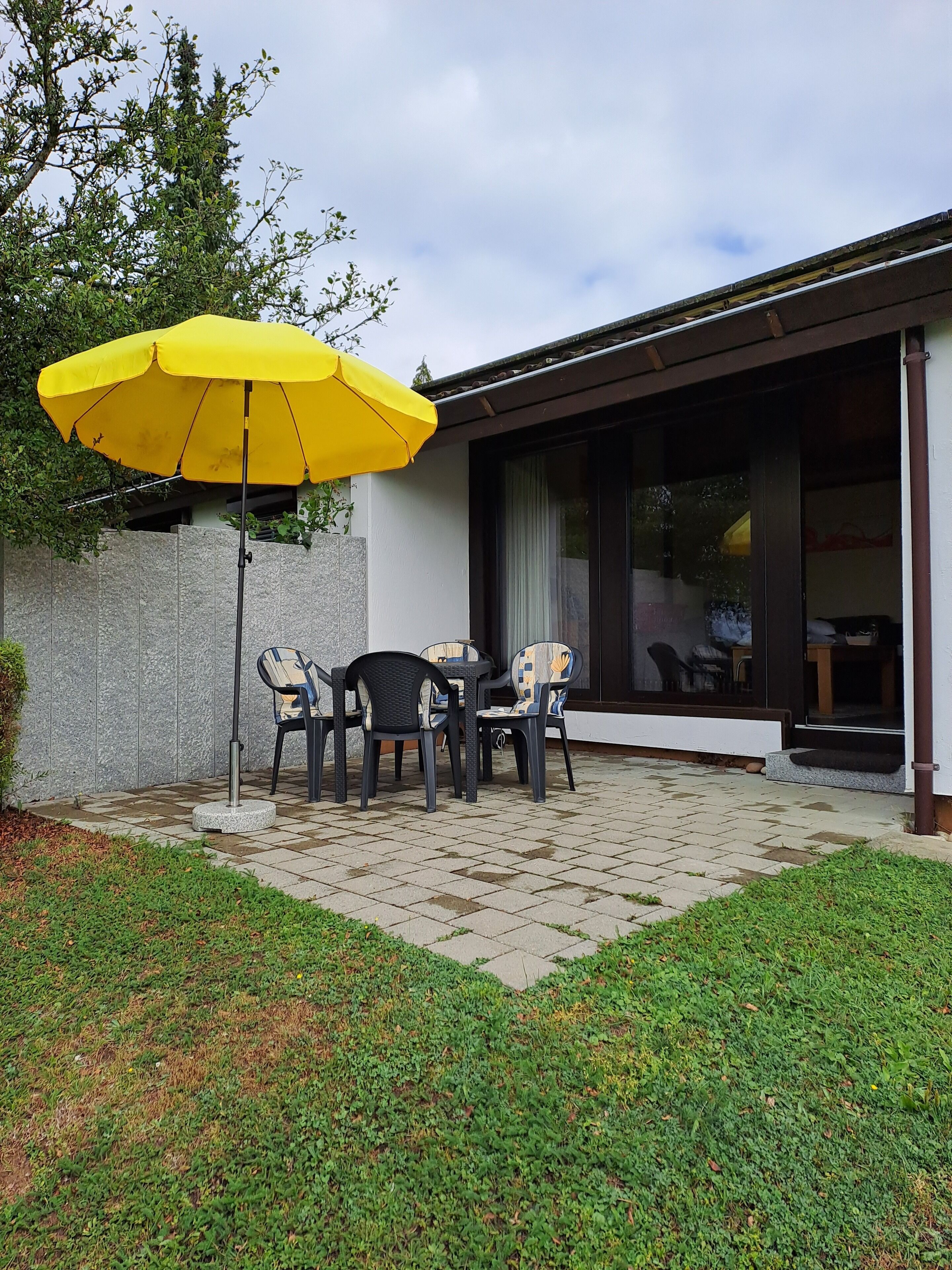 Un espace extérieur aménagé en terrasse, avec un parasol jaune, un sol carrelé en pierre et un ensemble de quatre chaises.