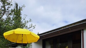 Ein Patio im Freien mit einem gelben Sonnenschirm, einem Boden aus Steinplatten und einer Sitzgruppe aus vier Stühlen.