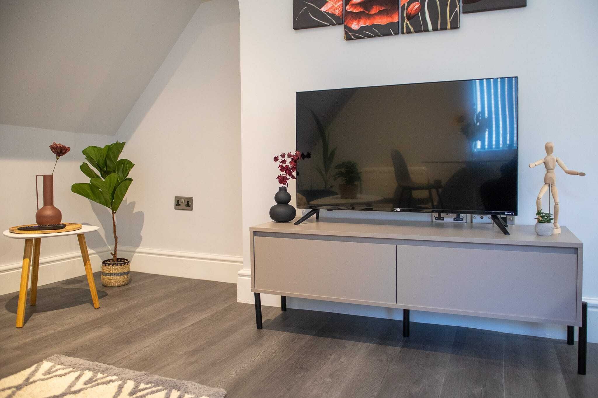 Una sala de estar moderna con un televisor de pantalla plana grande sobre una consola gris, una mesita redonda con un jarrón y una planta, y una planta verde grande.