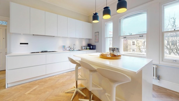 Une cuisine moderne avec des meubles blancs, un îlot central doté de tabourets de bar, un éclairage suspendu et de grandes fenêtres qui laissent entrer la lumière naturelle.