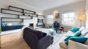 Un salon moderne avec un canapé foncé, un canapé de couleur claire agrémenté de coussins turquoise, une table basse en verre et une bibliothèque contenant divers objets.