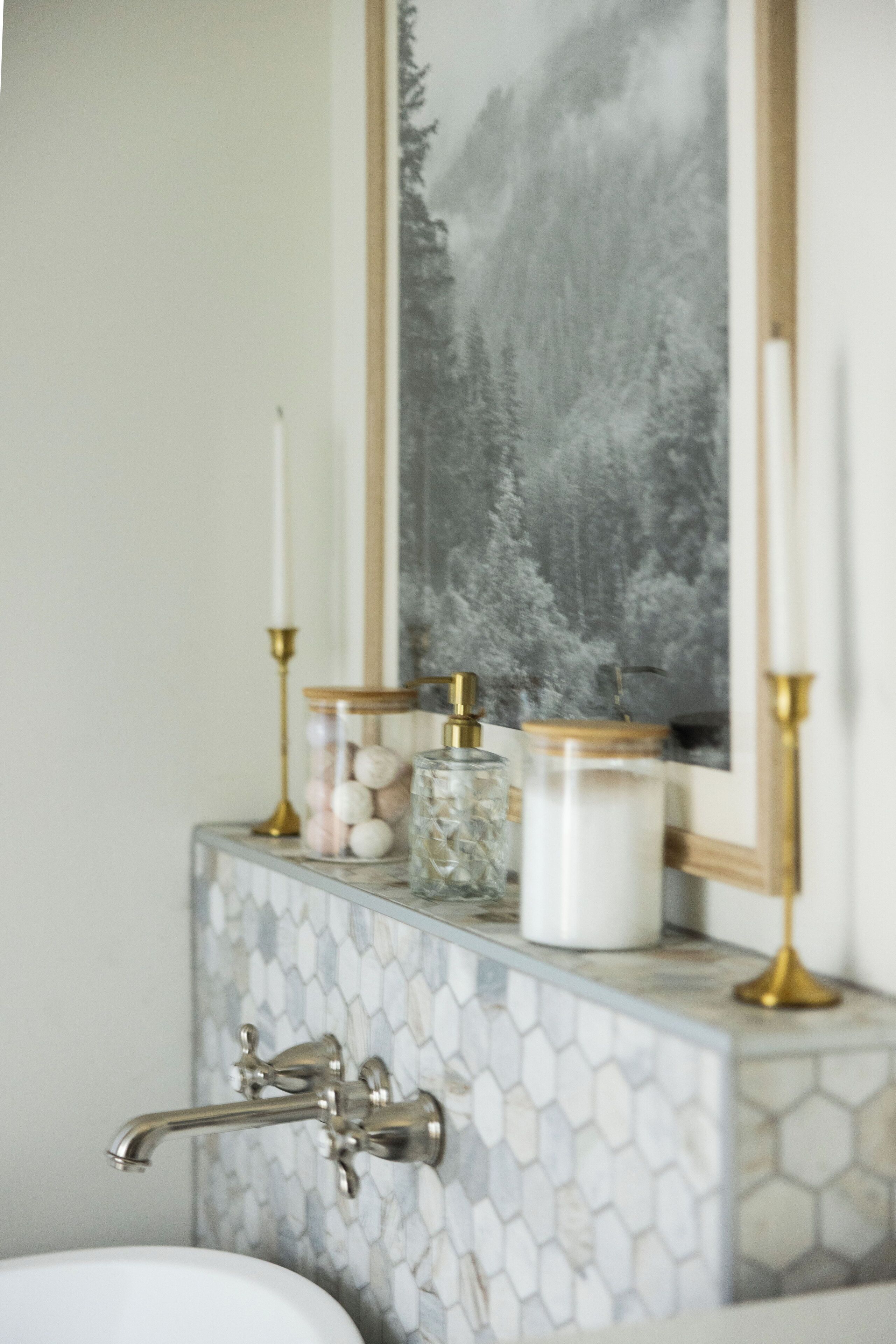 A bathroom vanity with a marble tiled backsplash, a gold faucet, and decorative items including a candle holder, jars, and a framed picture.