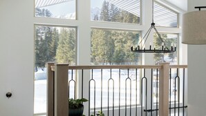 A spacious room with a wooden floor, a view of a snowy landscape through large windows, a hanging light fixture, and a chair with a cushion. - Modern Chalet with Spa Bathoom (Seward)