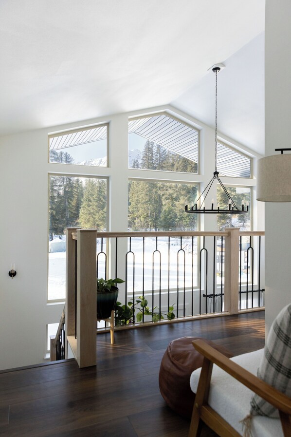 A spacious room with a wooden floor, a view of a snowy landscape through large windows, a hanging light fixture, and a chair with a cushion.