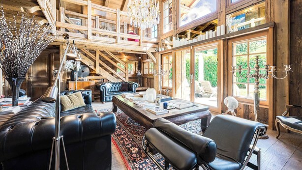 A spacious living area with a high ceiling, wooden beams, a chandelier, and a large window offering a view of greenery. - Luxury Stay - Comfortable Chalet With Jacuzzi (Megeve)
