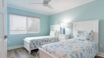 A bedroom with two beds, a ceiling fan, a window with blinds, and a bedside table with a lamp.