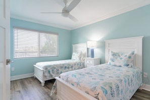 A bedroom with two beds, a ceiling fan, a window with blinds, and a bedside table with a lamp.