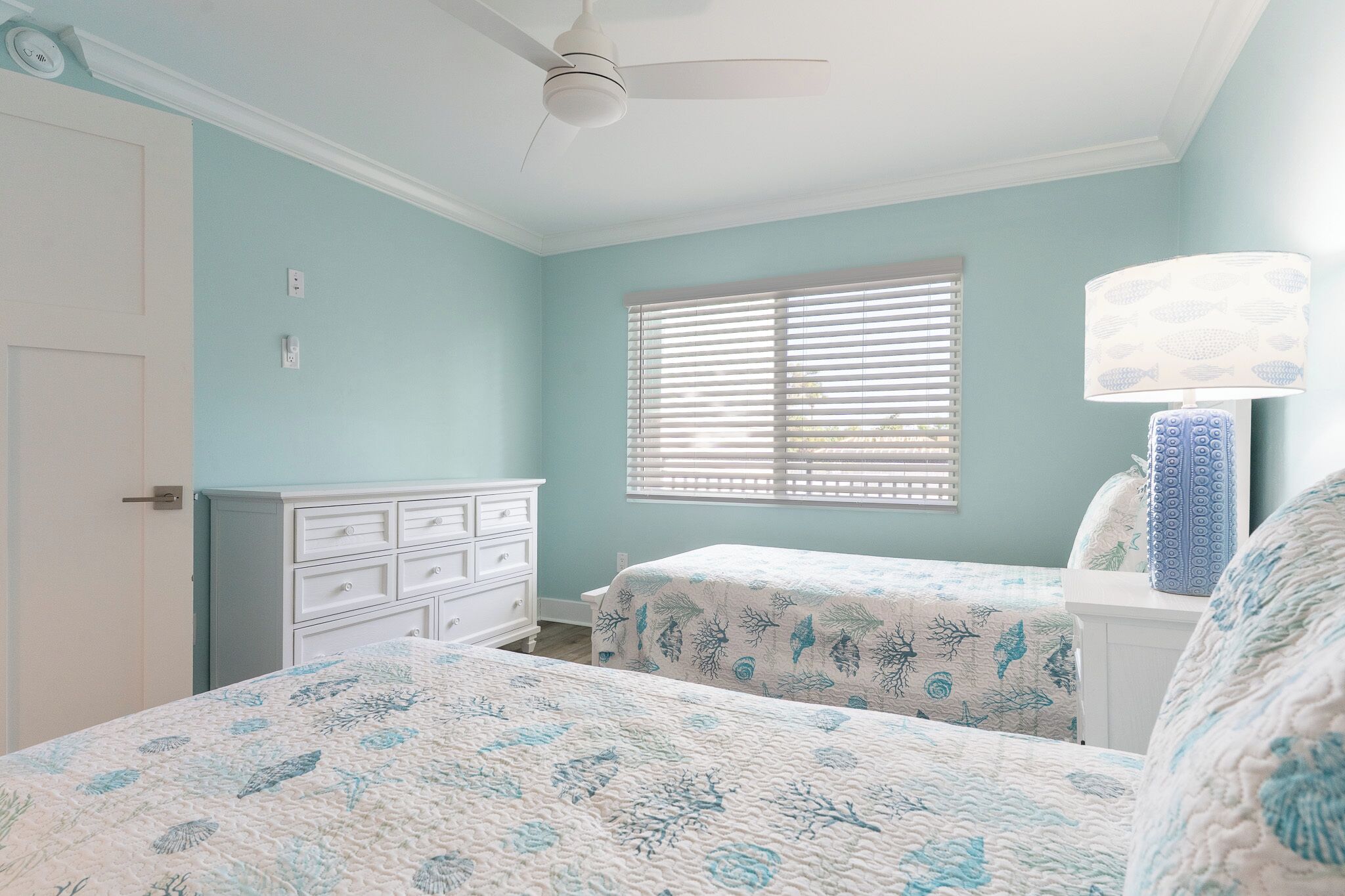 Une chambre avec deux lits, une commode, un ventilateur de plafond, une fenêtre avec des stores et une lampe.
