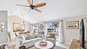 Un salon spacieux doté d’un ventilateur de plafond, d’une grande fenêtre avec des rideaux, d’un ensemble de canapés, d’une table basse et d’un téléviseur à écran plat.