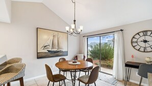 Un coin repas avec une table ronde, quatre chaises, un lustre, un grand tableau représentant un voilier et une horloge murale.