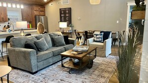 A cozy living room with a grey sofa, a coffee table, and a rug. There's a dining area with a table and chairs, and a kitchen in the background.