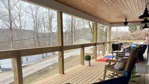 A covered deck with wooden flooring, a row of chairs, a table, and a view of a road and trees.