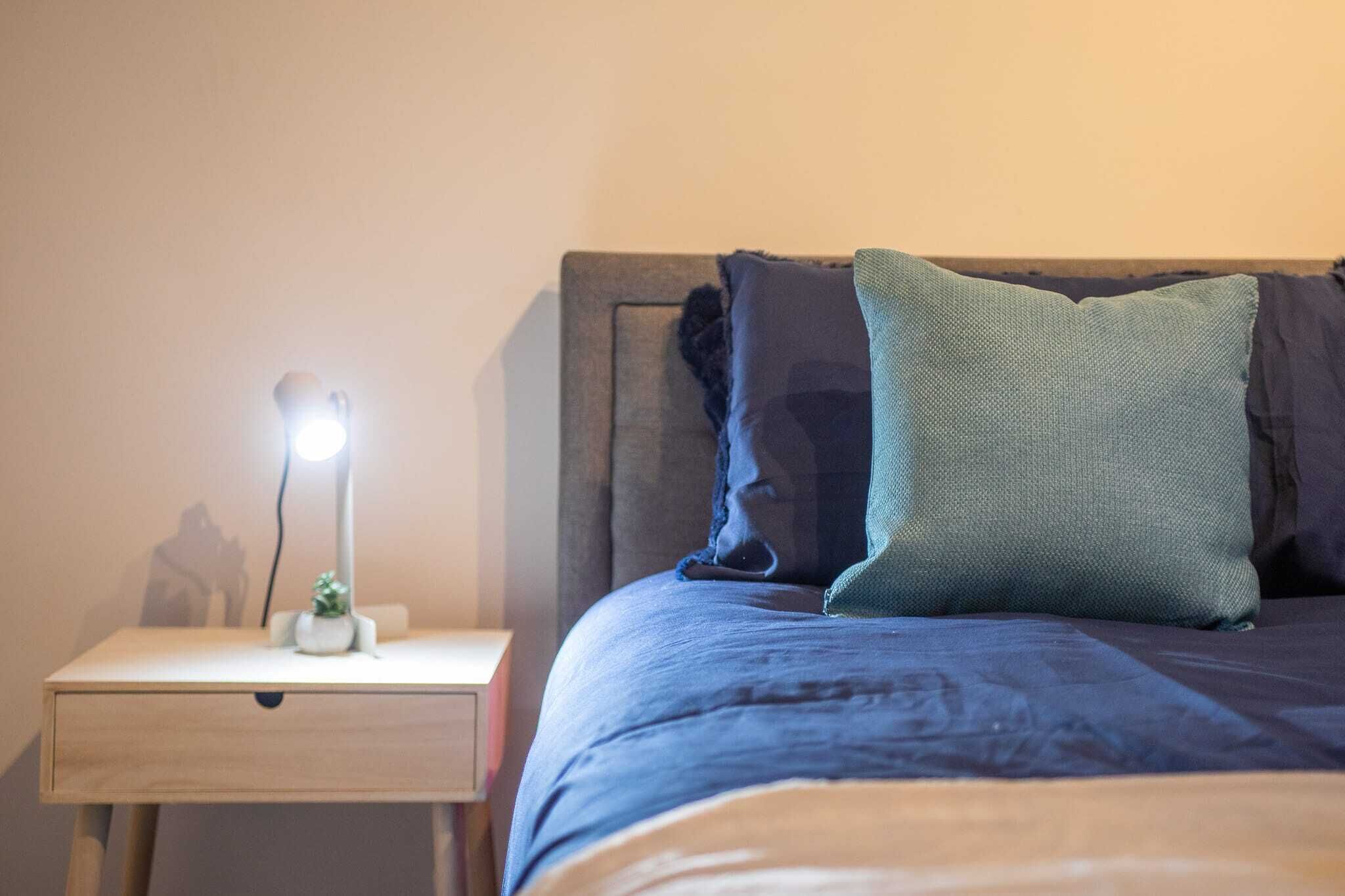 En pænt redt seng med et blåt tæppe og puder, et natbord med en lampe og en lille plante.