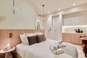 A modern hotel room with a large bed, a bedside table with a book, a small round table with a glass, and a kitchenette area. - Exceptional Grand Apartment - Louvre Museum (Paris)
