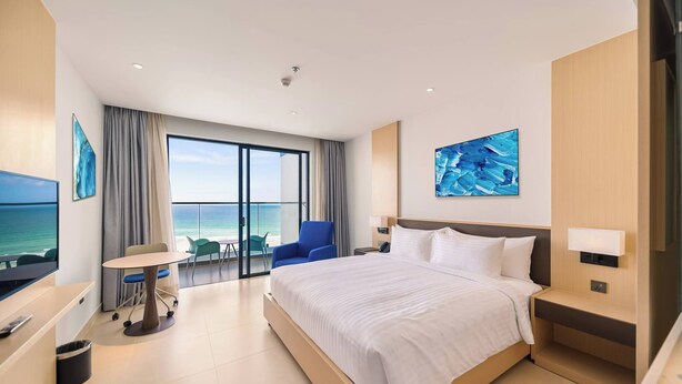 Una habitación de hotel moderna con una cama grande, un escritorio con silla, televisor y un balcón con vista al mar.