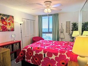 A bedroom with a bed, a red chair, a desk, a ceiling fan, a window with blinds, a painting, and a wall with a sign. - 908 Dover - Sea Colony East (Bethany Beach)