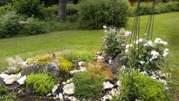 Un jardin où s’épanouissent une grande variété de plantes et de fleurs, notamment des roses blanches, le tout entouré de rochers et de verdure.