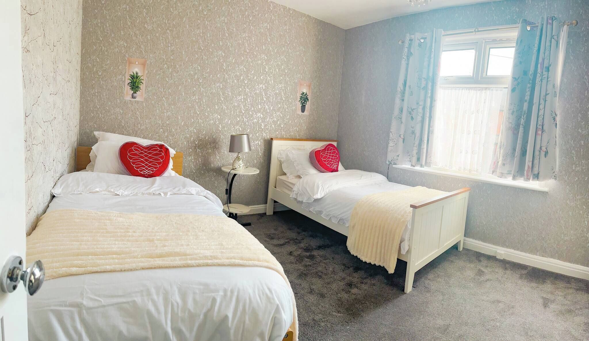 A bedroom with two single beds, a bedside table with a lamp, a small plant, and a window with curtains.