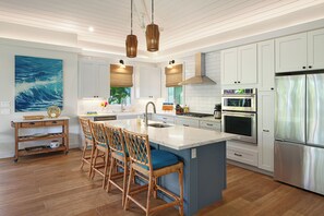 A modern kitchen with white cabinetry, a large island with bar stools, stainless steel appliances, and a ceiling fan. - Spacious Poipu Home with Guest House, Resort-Style Outdoor Living & Pool! (Koloa)