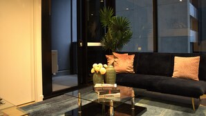 Uma sala de estar moderna com uma mesa de centro de vidro, um sofá preto com almofadas e uma grande janela com vista para a cidade.