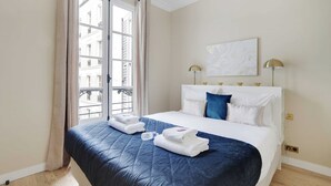 Et hotelværelse med seng, et vindue med gardiner, et billede på væggen og en lampe.