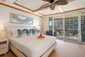 A bedroom with a large bed, a ceiling fan, a balcony with a view of the building, and a framed artwork on the wall.