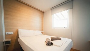 Uma cama de solteiro com roupa de cama branca, cabeceira de madeira, janela com persianas e uma tomada na parede.