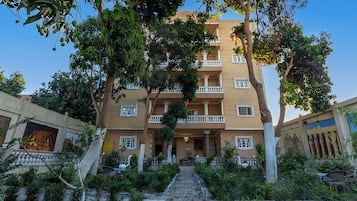 Un edificio de varios pisos con una entrada central, rodeado de vegetación y un sendero pavimentado que conduce a la entrada.