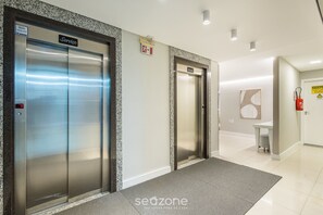 A modern elevator area with two elevators, a fire exit sign, and a small table with a chair in the background. - 2 Bdrm apt 250m From the Beach in Center Sdc0601 (Balneário Camboriú)
