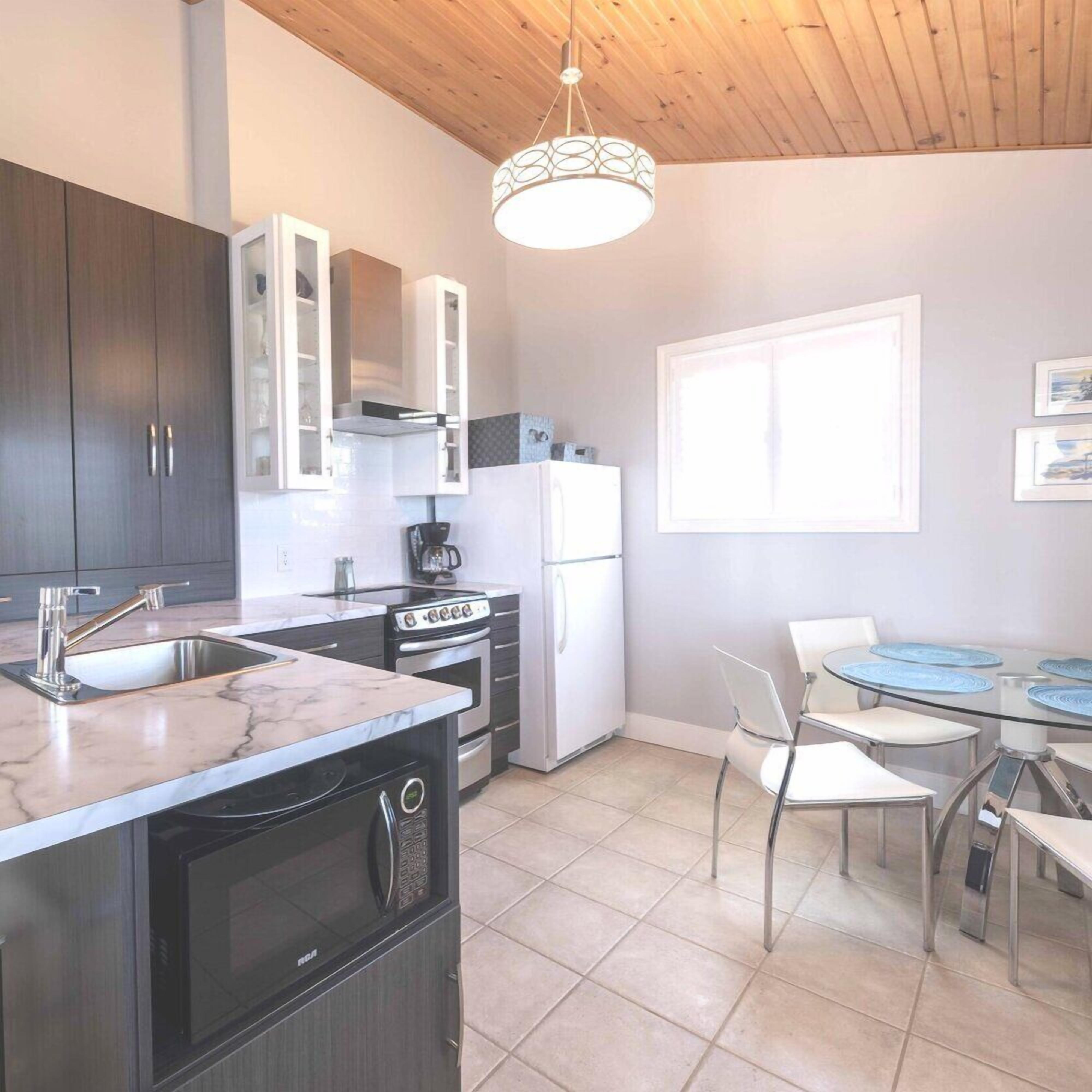 A modern kitchen with dark cabinets, a white refrigerator, a stove, and a sink. There is a dining area with a glass table and chairs.