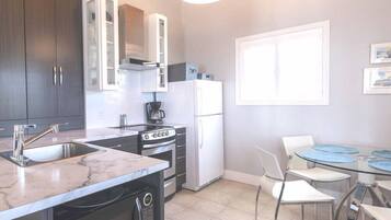 A modern kitchen with dark cabinets, a white refrigerator, a stove, and a sink. There is a dining area with a glass table and chairs.