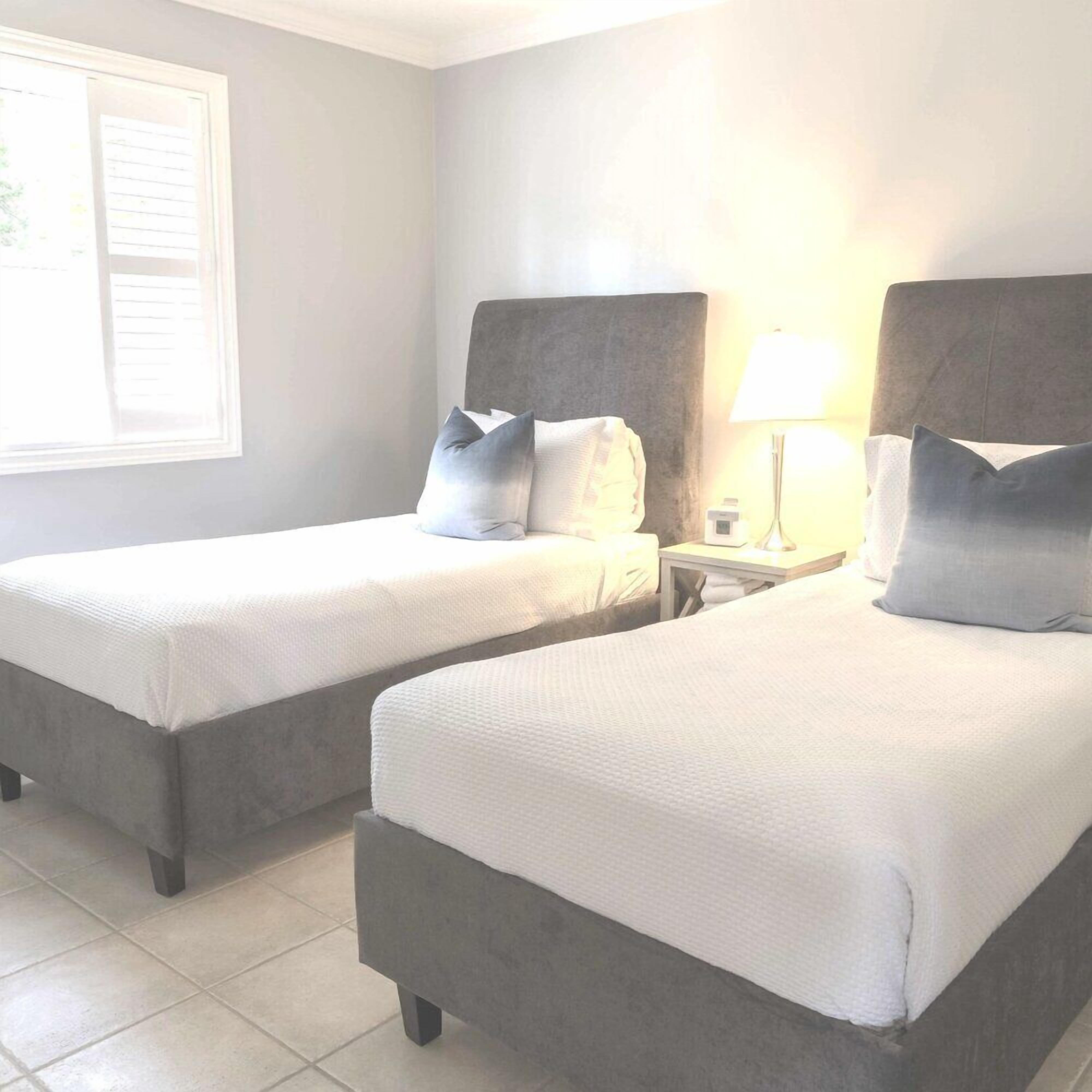 Een hotelkamer met twee bedden, elk met een grijs hoofdeinde, wit beddengoed en een nachtkastje met een lamp.