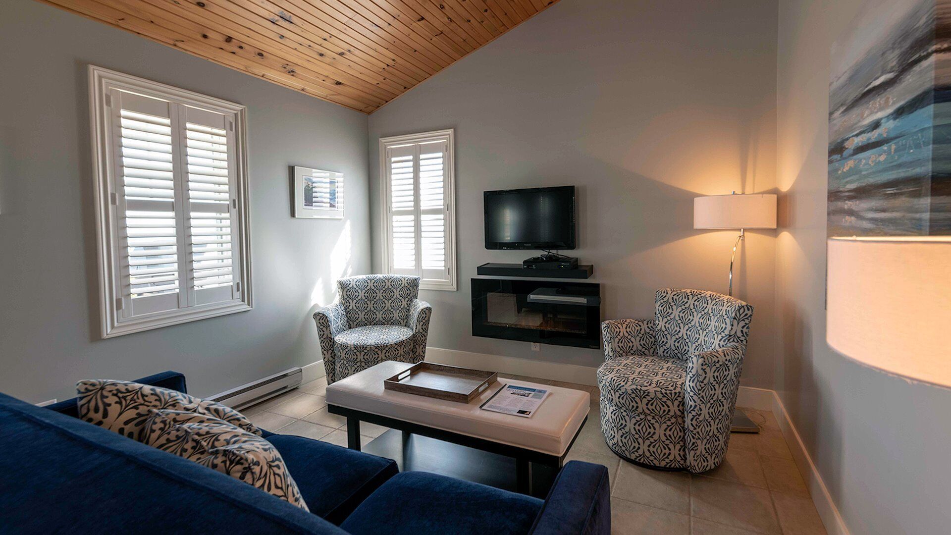 Ein Wohnzimmer mit einem blauen Sofa, gemusterten Sesseln, einem Couchtisch mit Büchern, einem Fernseher und einer Lampe.