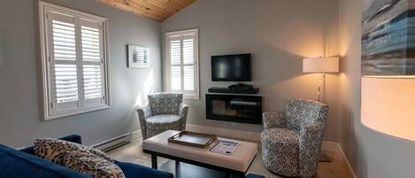 Ein Wohnzimmer mit einem blauen Sofa, gemusterten Sesseln, einem Couchtisch mit Büchern, einem Fernseher und einer Lampe.