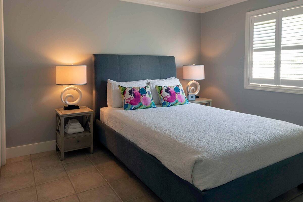 A bedroom with a bed, two bedside tables, a window with blinds, and a wall with a light switch.