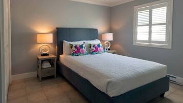 A bedroom with a bed, two bedside tables, a window with blinds, and a wall with a light switch.