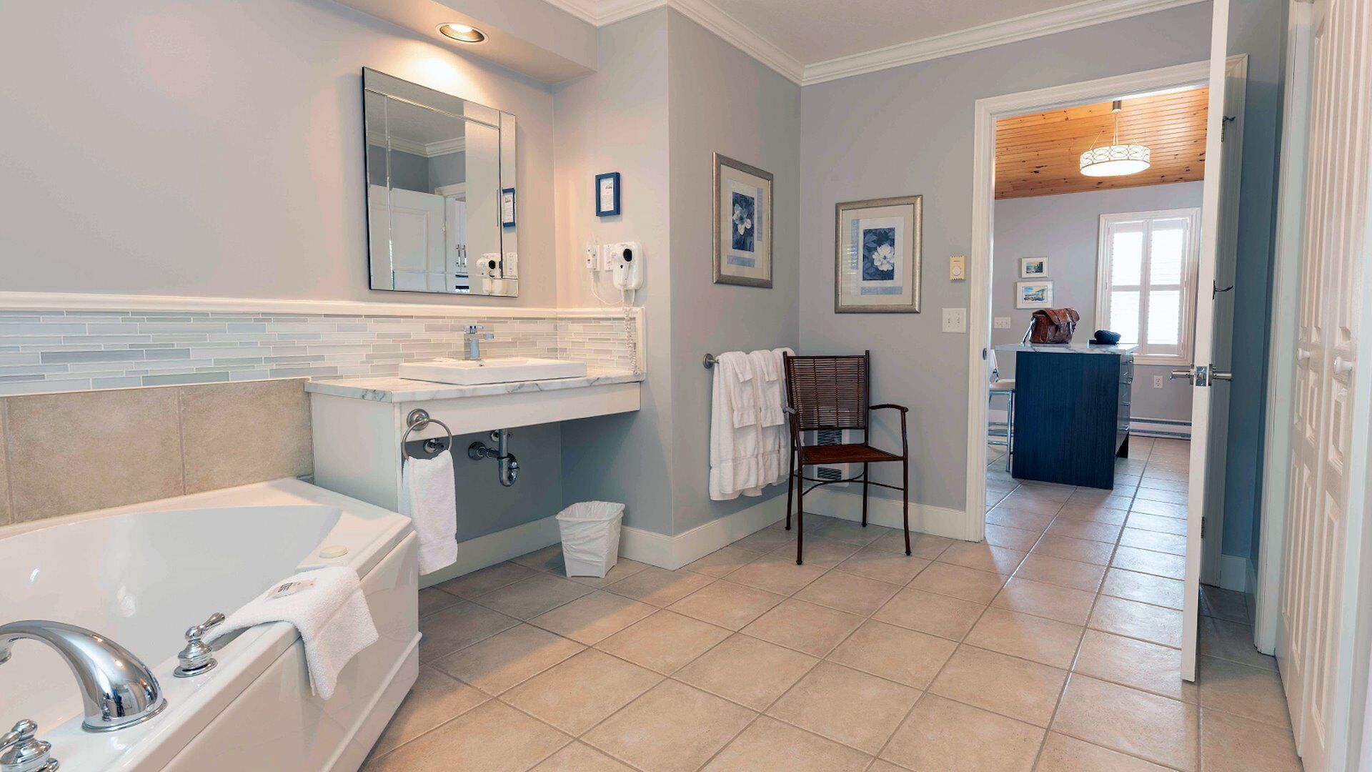 A bathroom with a bathtub, sink, and mirror. There is a chair and a trash bin.