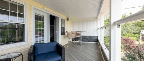 A covered porch with a wicker chair and a table, a door leading inside, and a view of greenery outside.