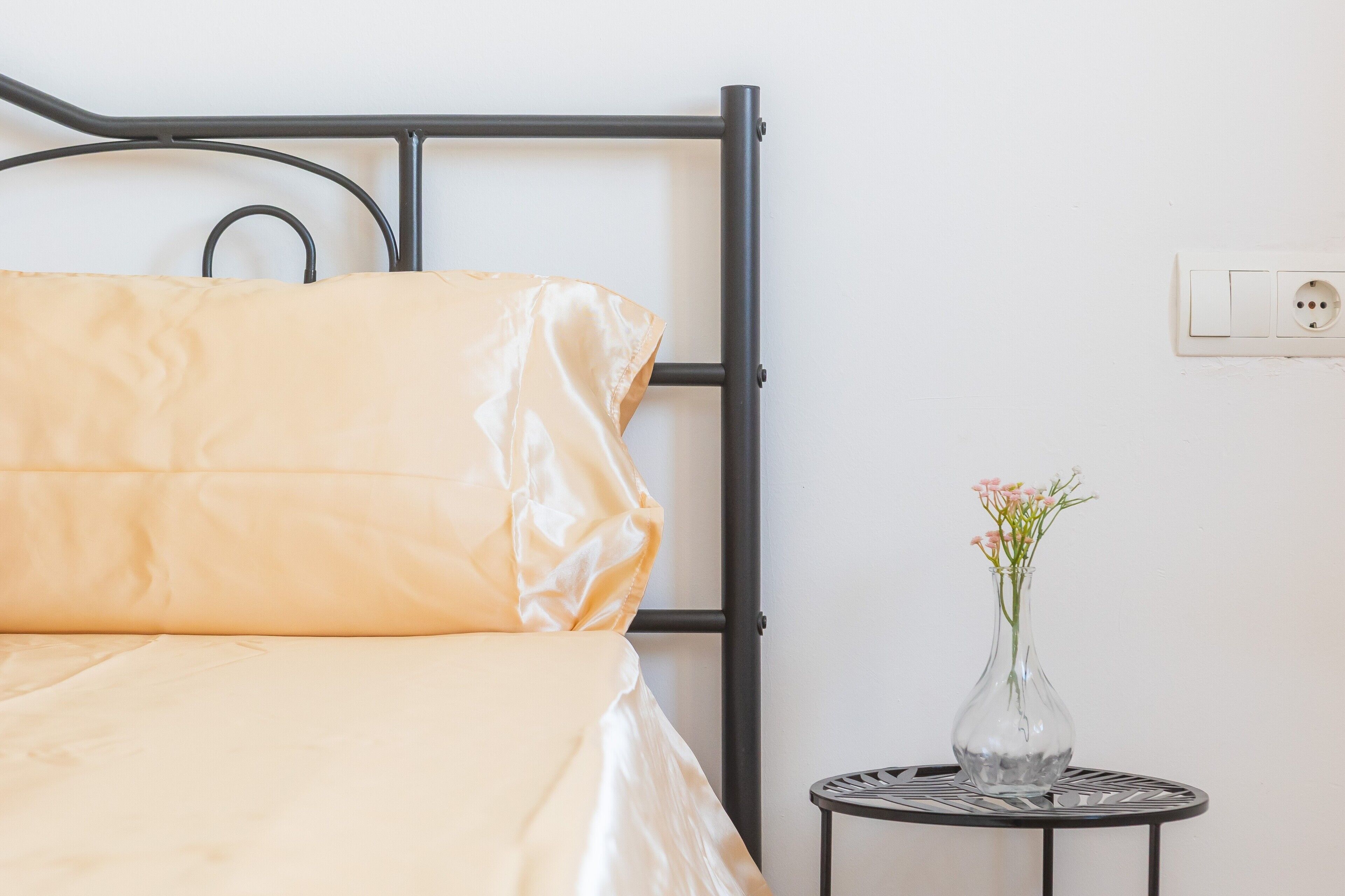 A bed with a metal frame, a small round table with a vase of flowers, and a power outlet on the wall.
