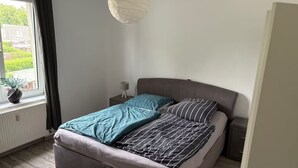 Une chambre à coucher comprenant un lit, une table de chevet, une lampe, une fenêtre avec des rideaux et une vue sur la verdure à l’extérieur.
