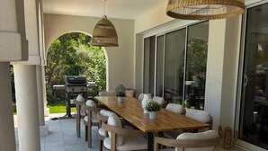 Eine überdachte Terrasse mit Holztisch und Stühlen, Korbschwinglampen und einem Grill.