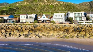 A coastal property with modern architecture, a stone retaining wall, and a view of the beach and hills. - 25% Off Sept 7-9, On The Sand Perfect 4 Families! (Ventura)