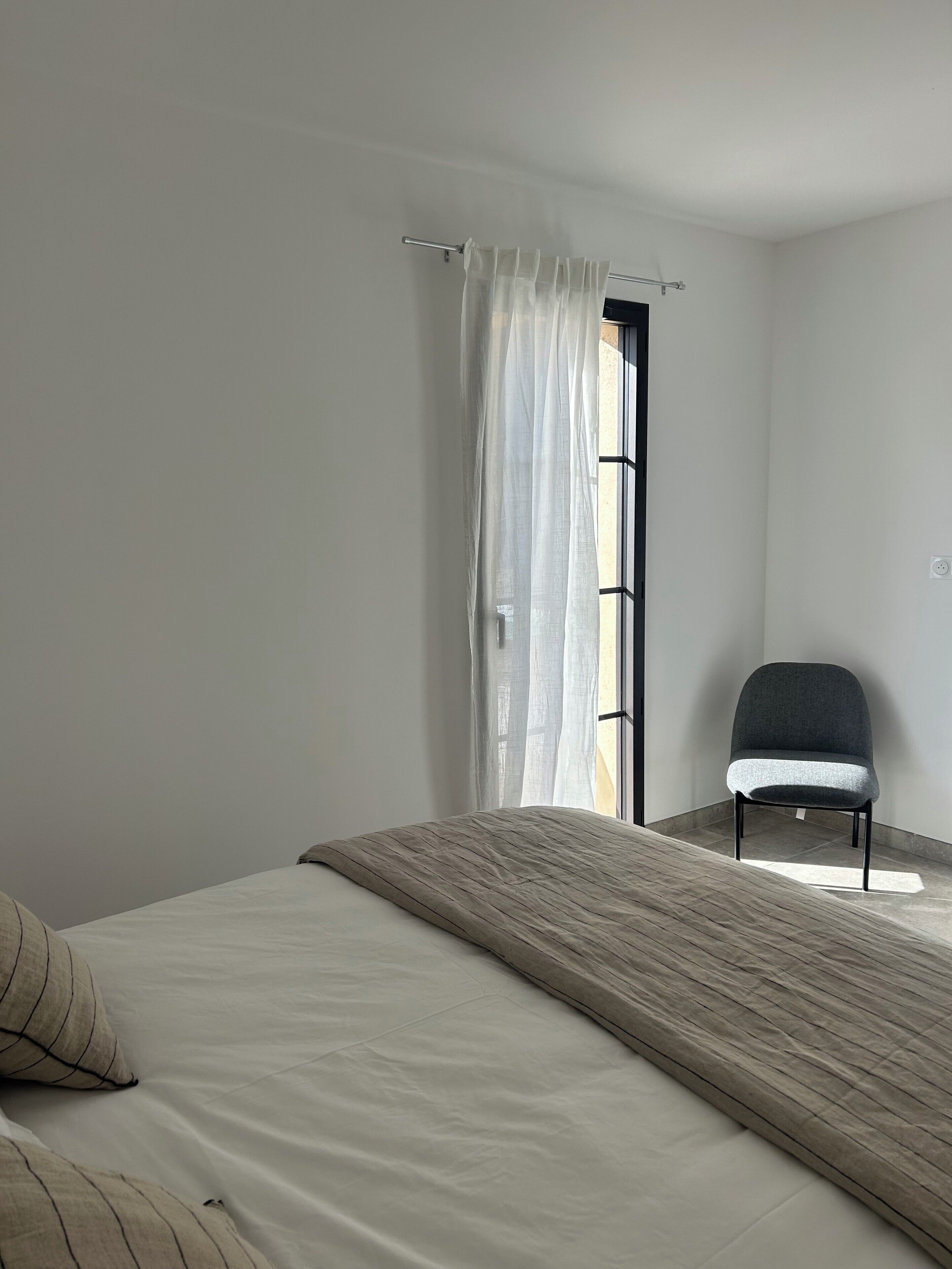 A minimalist bedroom with a bed, a chair, and a window with sheer curtains.